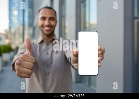 Junger, erfolgreicher Mann, der den weißen Bildschirm vor der Kamera zeigt, Geschäftsmann, der App empfiehlt, glücklich in die Kamera blickt, vor dem Bürogebäude. Stockfoto