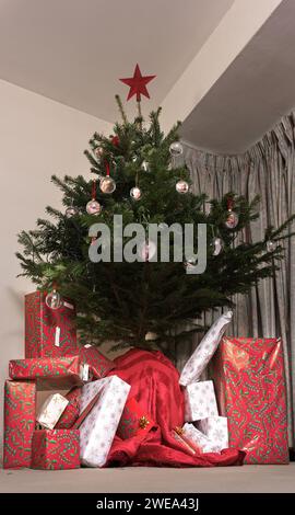 Weihnachtsgeschenke in buntes Papier gestapelt unter einem mit Kugeln geschmückten Weihnachtsbaum, der mit einem roten Stern gekrönt ist. Stockfoto