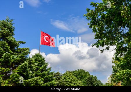 Türkische Flagge winkt im Wind vor einem blauen Himmel mit Wolken, umgeben von grünen, üppigen Bäumen Stockfoto