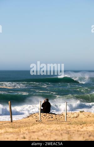 Ein älterer Mann am Telefon sitzt auf einer Sanddüne vor dem Meer. Stockfoto