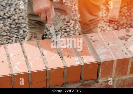 Nahaufnahme einer Mauer aus Ziegelstein und einer Kelle, die der Arbeiter zum Auftragen und Glätten des Mörtels zwischen Ziegelsteinen verwendet. Maurer, der den letzten Schliff auf den Ziegelstein legt Stockfoto