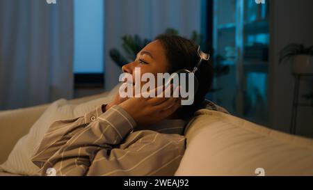 Entspannte afroamerikanische Frau in den Kopfhörern fallen auf bequemes gemütliches Sofa Abend Freizeit zu Hause Musik hören ethnische Mädchen Ruhe mit Augen Stockfoto