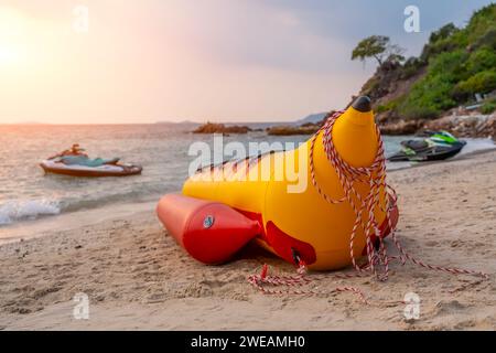 Aufblasbare Banane für reitende Touristen Menschen an einem sandigen malerischen tropischen Strand während Sonnenuntergang. Ein Jet-Roller im Wasser im Hintergrund. Stockfoto