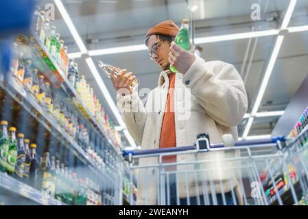 Der Mann im Laden wählt einen Drink aus. Der Typ im Supermarkt liest das Etikett, die Komposition von Limonaden, wählt, vergleicht Preise, sucht nach Vorteilen. Ansicht von unten vom Einkaufswagen. Regale mit einer großen Auswahl an Trinkwaren Stockfoto