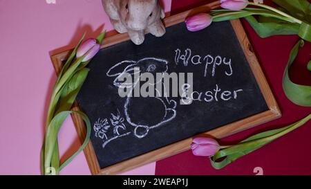 Eine Tafel mit einem osterhasen und frohe Ostern drauf. Pinkfarbene Tulpen. Rosa, lila Hintergrund. Urlaubskonzept. Stockfoto