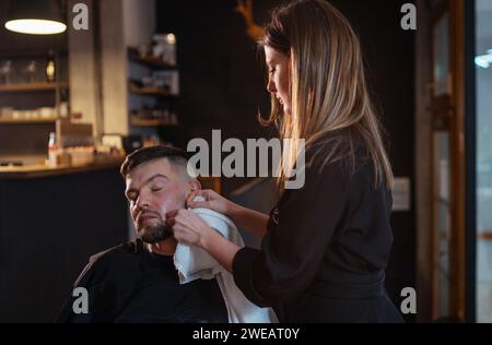 Langhaarige junge Barbierin rasiert sich sanft bärtige Männergesicht Borsten, während er im Stuhl sitzt. Sie entfernt Creme-Schaum-Reste von den Wangen. Haar und Haut c Stockfoto