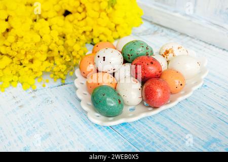 ostereier mit Mimosa. Osterferien Mimosenblumen dekoriert mit bunten Eiern. Schokoladenbonbons mit Zuckerguss in Form von Wachteleiern auf einem Arbeitsauftrag Stockfoto