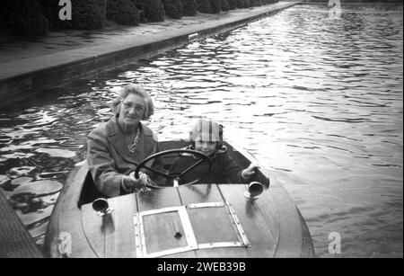 1950er Jahre, historisch, auf der Suche nach einem Foto, eine Dame und ein junges Mädchen sitzen in einem kleinen, hölzernen motorisierten Boot für zwei Personen auf einem von Bäumen gesäumten See oder Teich, England, Großbritannien. Stockfoto