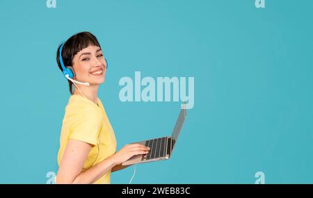 Lächelnde Frau in einem gelben T-Shirt und Kopfhörern mit Laptop, pc-Computer auf blauem Hintergrund mit Kopierraum. Arbeit Lernen, virtuelle Schulung, E-Lear Stockfoto