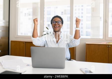 Überglücklich aufgeregte afroamerikanerin feiert den Sieg, sitzt im Coworking-Raum vor dem Laptop, glückliche Geschäftsfrau schrie ja, hat gute Nachrichten zurückgeholt, Stellenangebot Stockfoto