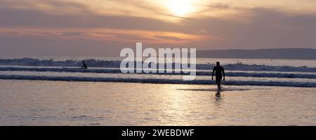 Ein Mann in Silhouette geht vom Wasser weg, während ein Surfer die Wellen bei Sonnenuntergang mit der Insel dahinter in Essaouira, Marokko, am 24. Januar 2024 angreift Stockfoto