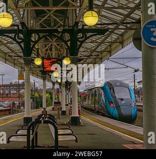 Ein Zug fährt von einem Bahnhof in einem Abendlicht ab. Lampen unter einem Vordach aus dem 19. Jahrhundert sind beleuchtet, und im Vordergrund befindet sich eine Bank. Stockfoto