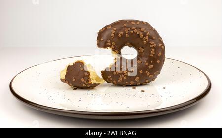 Ein Schokoladenkrapfen mit Streuseln auf einem weißen Teller. Ein abgebissener Teil des Donuts, der daneben lag, als hätte jemand ein Stück abgebissen. Stockfoto