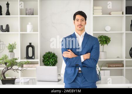 Ein zuversichtlicher hispanischer Geschäftsmann steht mit überkreuzten Armen in einem modernen Büro, seine ausgeglichene Haltung ergänzt das elegante, moderne Büro. Professionalität und Entschlossenheit Stockfoto