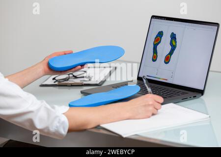 Orthopädie, die in einer Klinik speziell angefertigte Einlegesohlen vor dem Laptop hält, mit einem Testfoto. Fußentspannung und orthotische Medi Stockfoto