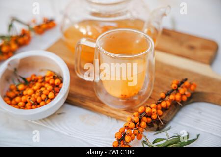 Tasse mit duftendem Vitamintee aus Sanddorn auf einem Holztisch. Frische Sanddornbeeren auf einem Zweig sind bereit für die Vorbereitung einer Heilung Stockfoto
