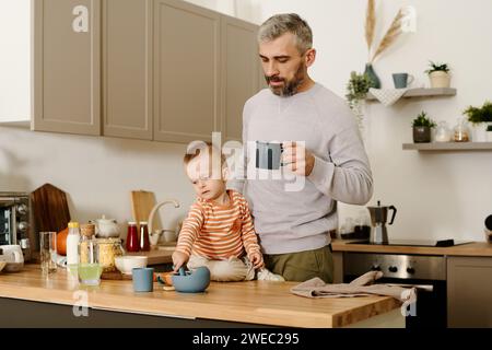 Reifer Vater mit einer Tasse Tee oder Kaffee, der neben seinem kleinen Sohn sitzt und am Küchentisch mit Küchenutensilien beim Frühstück sitzt Stockfoto