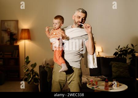 Reifer bärtiger Vater und sein liebenswerter Sohn putzen bei der Morgenhygiene im Wohnzimmer Zähne mit Zahnbürsten Stockfoto