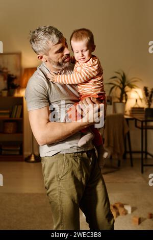 Reifer Mann in grauem T-Shirt und beigefarbener Hose, der einen schreienden, frechen Sohn an den Händen hält und ihn ansieht, während er im Wohnzimmer steht Stockfoto