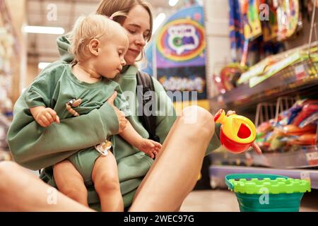 Junge attraktive Frau mit ihrem süßen Baby, das auf dem Boden im Spielwarenladen sitzt. Wunderschöne Mutter und ihre kleine blonde Tochter, die im Einkaufszentrum einkaufen und hav Stockfoto