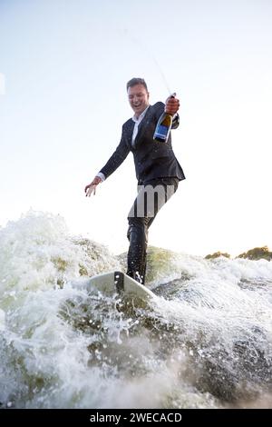 Junger Mann im klassischen Anzug mit Champagnerflasche fährt auf einem Wakeboard auf dem See in der Nähe der Stadt. Happy Clerk ist aus einem stickigen Büro geflohen, um aktive sp Stockfoto