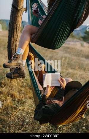 Ästhetische Ruhe. Ein wunderschönes Mädchen und ihr Freund entspannen sich bei Sonnenuntergang in Hängematten. Jugendliche lesen und spielen Musikinstrumente. Stockfoto