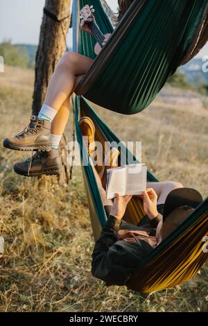 Ästhetische Ruhe. Ein wunderschönes Mädchen und ihr Freund entspannen sich bei Sonnenuntergang in Hängematten. Jugendliche lesen und spielen Musikinstrumente. Stockfoto