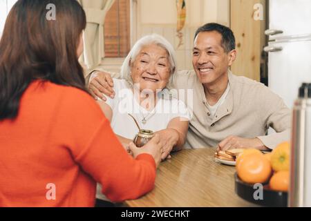 Glückliche Erwachsene Kinder teilen sich mit ihrer älteren japanischen Mutter, die in der Küche trinkt. Argentinisch-japanische Familie. Stockfoto