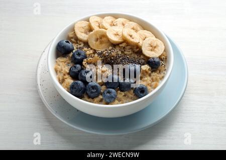 Leckeres Haferflocken mit Bananen, Heidelbeeren und Chiasamen, serviert in einer Schüssel auf weißem Holztisch Stockfoto