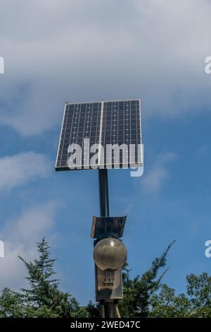 Kleines Solarmodul auf dem Lichtmast über Baumkronen vor blauem bewölktem Himmel Stockfoto