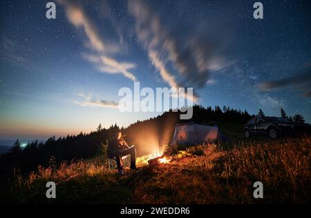 Camping in den Bergen unter bewölktem Nachthimmel. Ein junger Mann ruht sich am Lagerfeuer aus, in der Nähe eines Touristenzelts und eines Geländewagens auf einem Hügel. Ein reisender Mann, der nachts die Sterne beobachtet. Stockfoto