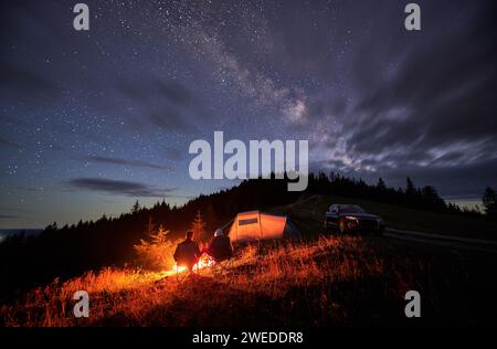 Glückliches Paar, das auf den Sternenhimmel mit Wolken starrt. Junge Menschen sitzen auf Stühlen und wärmen sich am Lagerfeuer in der Nähe von Touristenzelt und Geländewagen. Reisendes Paar, das sich zusammen ausruht. Camping bei Nacht. Stockfoto