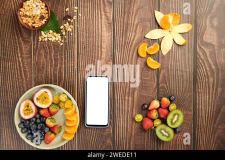Smartphone mit leerem Bildschirm auf Holztisch mit Obst und Müslischale zum Frühstück Stockfoto