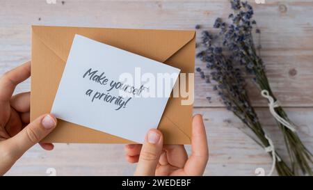 Weibliche Hände, die eine Notiz aus Papier mit Text nehmen, MACHEN SICH aus beigem Umschlag Eine PRIORITÄT. Lavendelblume. Draufsicht, flach. Konzept der psychisch-spirituellen Gesundheit Selbstversorgung Wohlbefinden Achtsamkeit Stockfoto