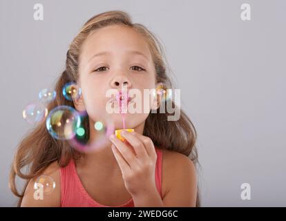 Porträt, kleines Kind und Blasen im Studio von Spielzeug, süße und lustige Spiele mit Seifenflüssigkeit. Mädchen, Gesicht und lernen, mit dem Blasstab zu spielen Stockfoto