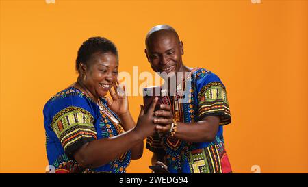 Traditionelles afroamerikanisches Paar, das Websites auf dem Smartphone durchsucht, Fotos in der Social-Media-App ansieht und über Memes lacht. Fröhliche Menschen in bunten Gewändern, die sich online Bilder anschauen. Stockfoto