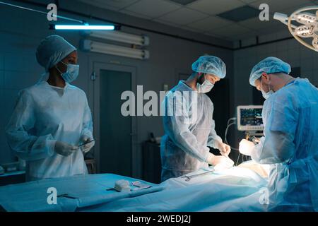 Multiethnisches Team von professionellen männlichen Chirurgen und weiblichen Krankenschwestern führt invasive Operationen am Patienten im Krankenhaus durch. Afroamerikanische Assistentin hilft Stockfoto