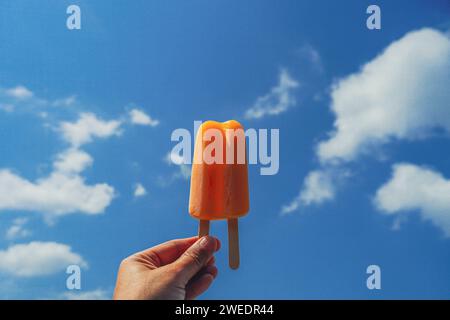 Orangeneis auf blauem Himmel Hintergrund. Orangenes Eis in der Frauenhand. Weibliche Hand, die schmelzendes oranges Eis auf blauem Himmel in Summe hält Stockfoto