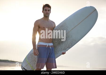 Surfen, Fitness und Porträt des Mannes am Strand mit Surfbrett für Spaß, Reisen und Sport im Freien. Ozean, Training und männliche Surfer am Meer für Stockfoto