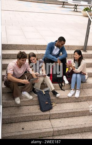 Multirassische junge Studenten, die auf Treppen sitzen. Verschiedene Gruppe glücklicher Leute, die draußen Akten lesen. Stockfoto