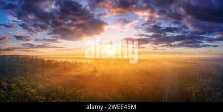 Nebeliger Herbstwald bei Sonnenaufgang aus der Vogelperspektive. Farbenfrohe Landschaft mit dramatischem Himmel, Morgennebel und dichtem Kiefernwald mit Eisenbahnpass Stockfoto
