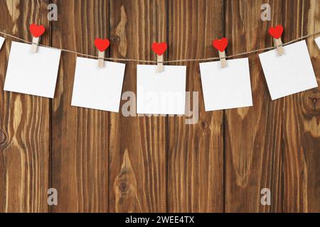 Kleiderschalen mit roten Herzen und leeren Blättern Papier auf Schnur vor braunem Holzhintergrund. Valentinstag, romantisches Konzept. Modell, tem Stockfoto