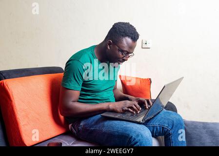 Junger Mann in Brille, der auf einem Sofa sitzt und mit Laptop arbeitet, lächelt. Stockfoto