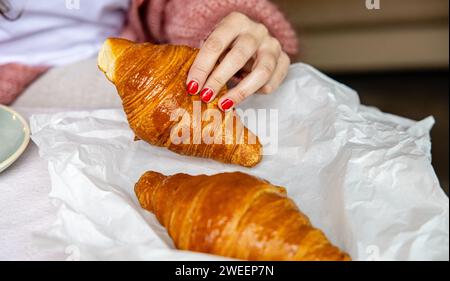 Frauenhand mit roten Nägeln, die ein goldgebackenes Croissant schnappt, symbolisiert einen frischen Start in den Morgen. Stockfoto