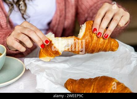 Nahaufnahme der Hände mit rotem nagellack, die ein Croissant brechen, ein Symbol für ein gemütliches Morgenmahl. Stockfoto