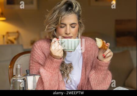 Eine zufriedene junge Frau in einem rosafarbenen Pullover genießt das Aroma ihres Kaffees mit geschlossenen Augen und hält ein frisch gebissenes Croissant. Stockfoto