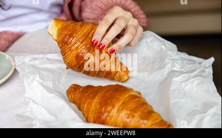 Frauenhand mit roten Nägeln, die ein goldgebackenes Croissant schnappt, symbolisiert einen frischen Start in den Morgen. Stockfoto