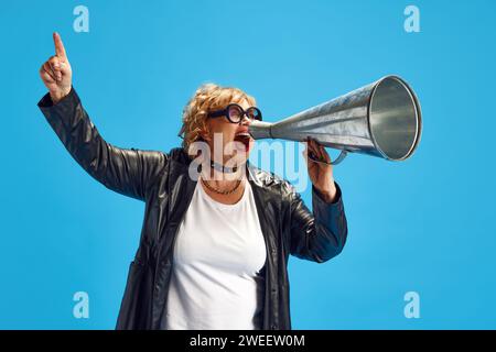 Frau, Rentnerin in Ledermantel gekleidet, die laut wichtige Neuigkeiten zum Retro-Megaphon vor blauem Hintergrund ruft. Stockfoto