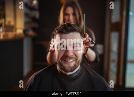 Lächelnder bärtiger Mann Porträt beim Friseurfriseur im Friseursalon von einer jungen Friseurfrau. Moderner Friseurladen in dunklem Schwarz Stockfoto