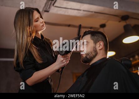 Junge langhaarige weibliche Friseur Friseur trocknende Haare mit professionellem Haartrockner bärtiger Mann im Salon Stuhl. Moderner Friseur im schwarzen Stil Stockfoto